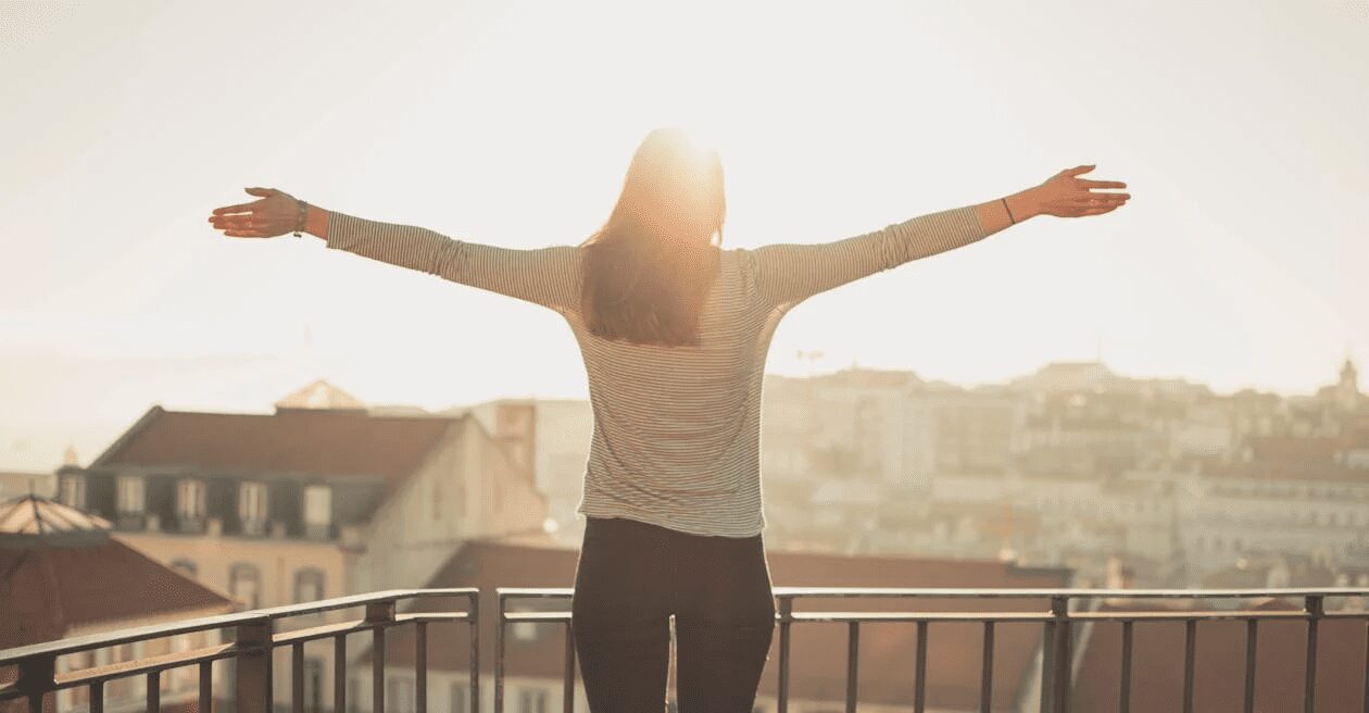 A woman with her arms outstretched, standing on a balcony with a city view in the background, illuminated by sunlight. 6 Empowering Ways to Reclaim How You Feel About Yourself