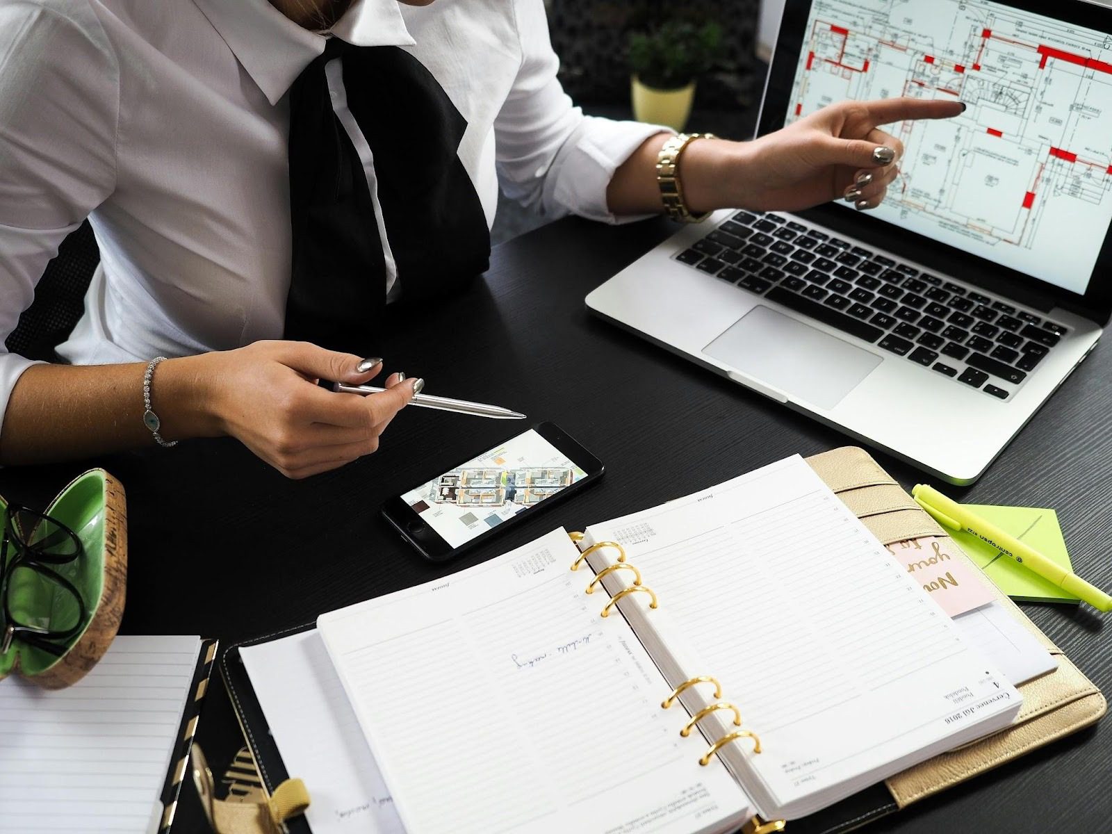 A person in a white shirt with a black bow tie holding a pen, discussing real estate plans using a laptop and smartphone, with a notebook and glasses on the desk. The Truth About Real Estate Investing: Profit, Pitfalls, and Smart Strategies