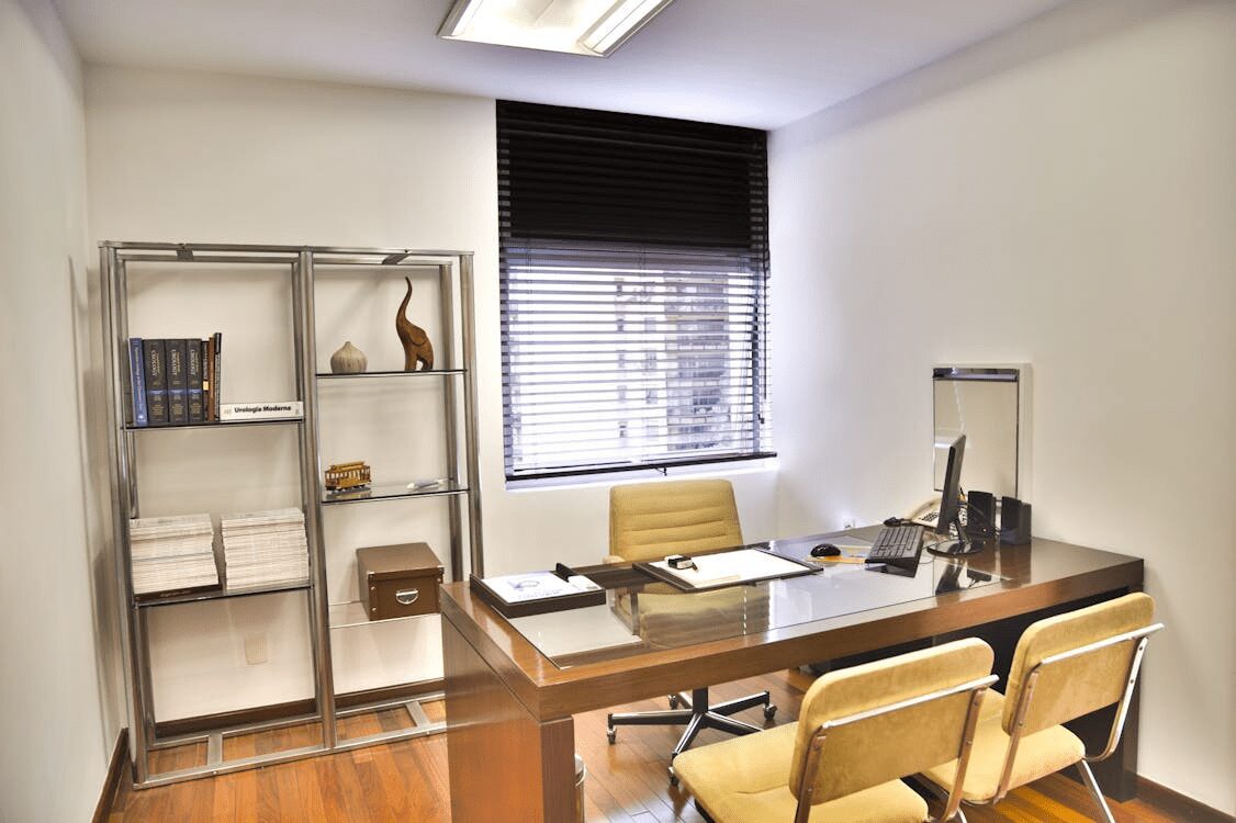 10 Ways to Make Clients Feel Comfortable in Your Office 7 A modern office setting featuring a wooden desk with a glass top, an ergonomic chair, and a shelving unit displaying books and decorative items. The room has ample natural light from a window with blinds. 10 Ways to Make Clients Feel Comfortable in Your Office