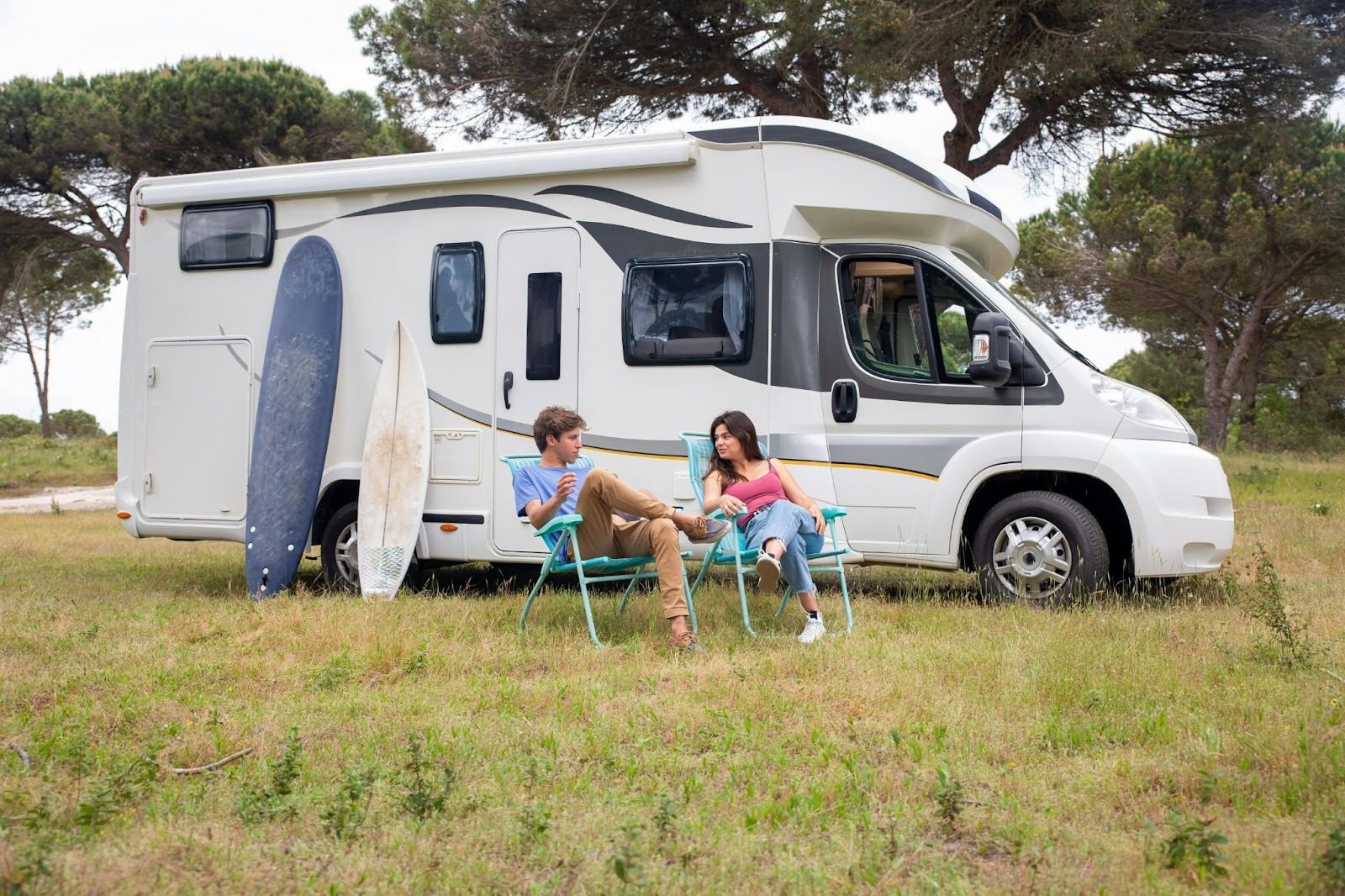 Road Trip Tips for Comfort and Adventure 7 A couple sitting on folding chairs beside a parked RV, with surfboards leaning against the vehicle, in a grassy field surrounded by trees. Road Trip Tips for Comfort and Adventure